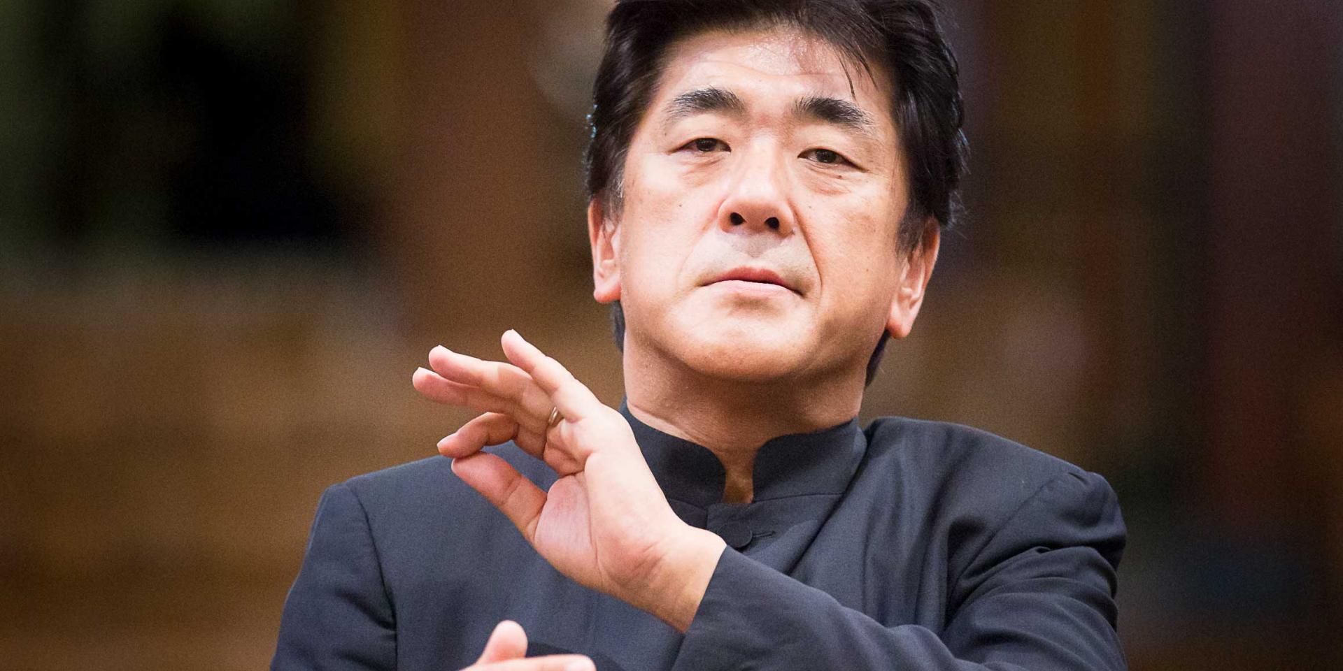 Yutaka Sado ripreso frontalmente mentre dirige, indossa una giacca grigio scuro alla coreana