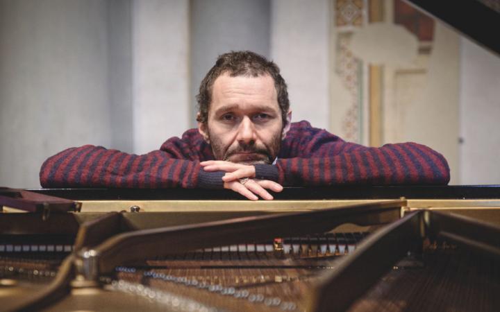 A front-facing portrait of musician Davide “Boosta” Dileo, leaning against the edge of an open grand piano.
