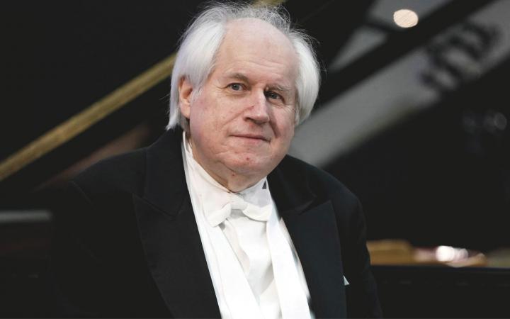 A waist-up portrait of pianist Grigory Sokolov with a grand piano in the background.