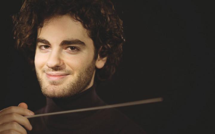A close-up portrait of conductor Emmanuel Tjeknavorian against a black background.