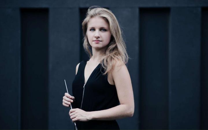A young blonde female conductor in a black dress holds her baton in front of her, against a dark grey panelled background