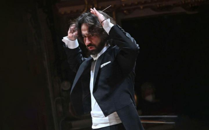 A dark-haired, bearded conductor in tailcoat directs with intense energy, raising his baton above his head.A dark-haired, bearded conductor in tailcoat directs with intense energy, raising his baton above his head