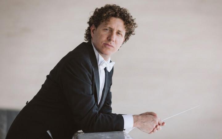 A curly-haired conductor in tailcoat and white bow tie, leaning on a surface, holds his baton while gazing thoughtfully at the camera