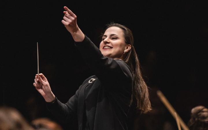 A young dark-haired female conductor in a black dress directs with a radiant smile, baton in hand and left arm raised,in front of a dimly lit orchestra
