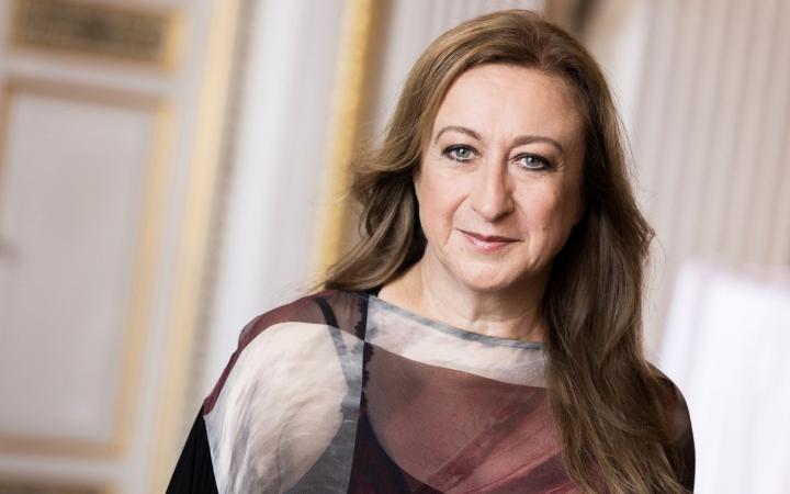 A female conductor with long brown hair and light eyes wears a geometric burgundy and grey blouse, portrayed with a serene expression in an elegant classical interior