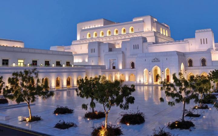 Illuminated facade of the Royal Opera House Muscat at dusk, a large white building in Omani style overlooking a paved square with trees.